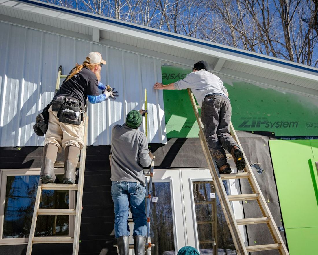 Three tradespeople on ladders installing siding and sheathing on a building exterior
