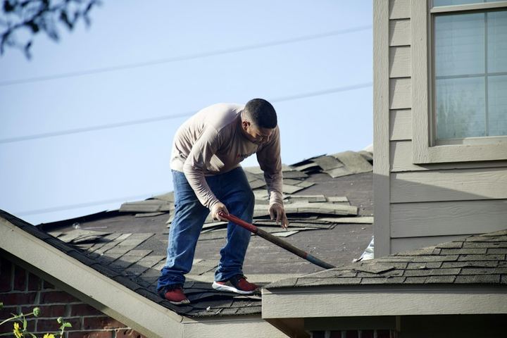 A worker tearing off damaged shingles from a house roof